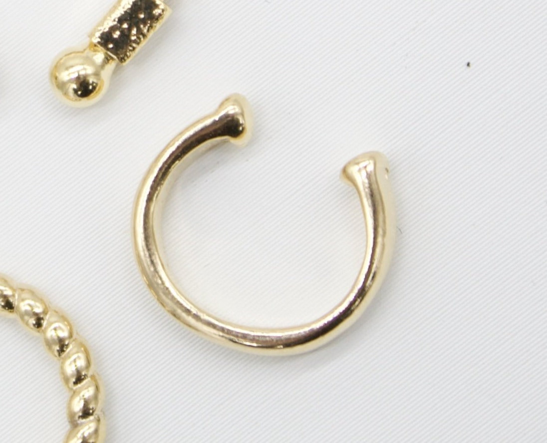 A minimalist gold ear cuff rests on a white background alongside other gold jewelry pieces.