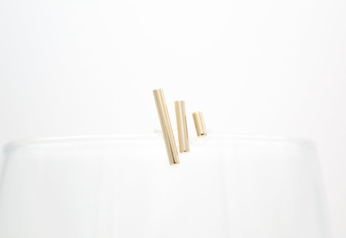Three minimalist gold bar earrings rest on the rim of a clear glass against a white background.