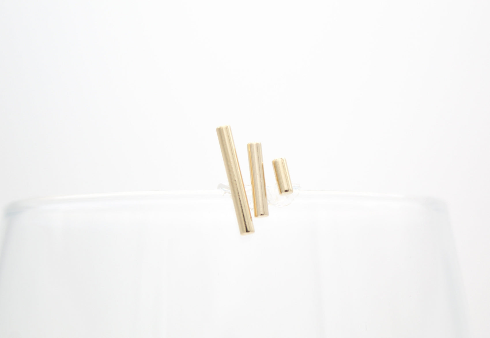 Three minimalist gold bar earrings rest on the rim of a clear glass against a white background.