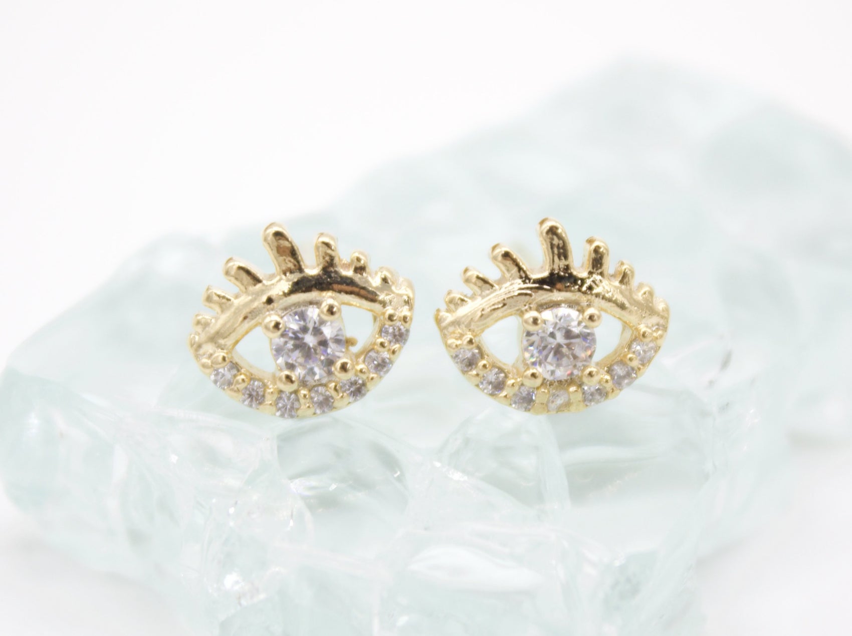 A pair of gold evil eye stud earrings featuring sparkling cubic zirconia crystals rests on a textured glass surface.