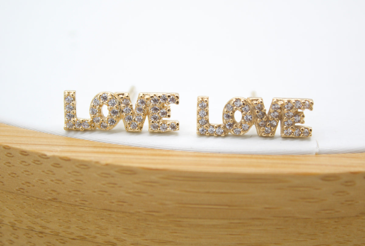 Two gold "LOVE" stud earrings encrusted with tiny sparkling diamonds rest side by side on a white surface behind a wooden display block.