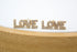 Two gold "LOVE" stud earrings encrusted with tiny sparkling diamonds rest side by side on a white surface behind a wooden display block.