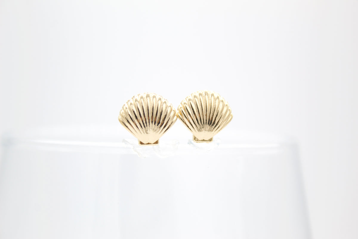 A pair of shiny gold seashell stud earrings rests on a glass surface against a clean white background.