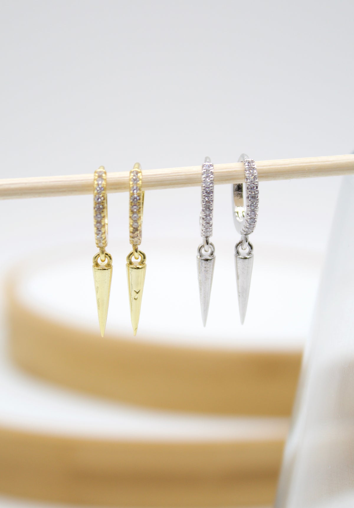 Two pairs of pave-set huggie hoop earrings with dangling spike charms hang from a wooden dowel, featuring gold-plated and silver-toned options.