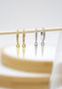 Two pairs of pavé huggie hoop earrings with dangling spike charms hang on a wooden stick against a white background.