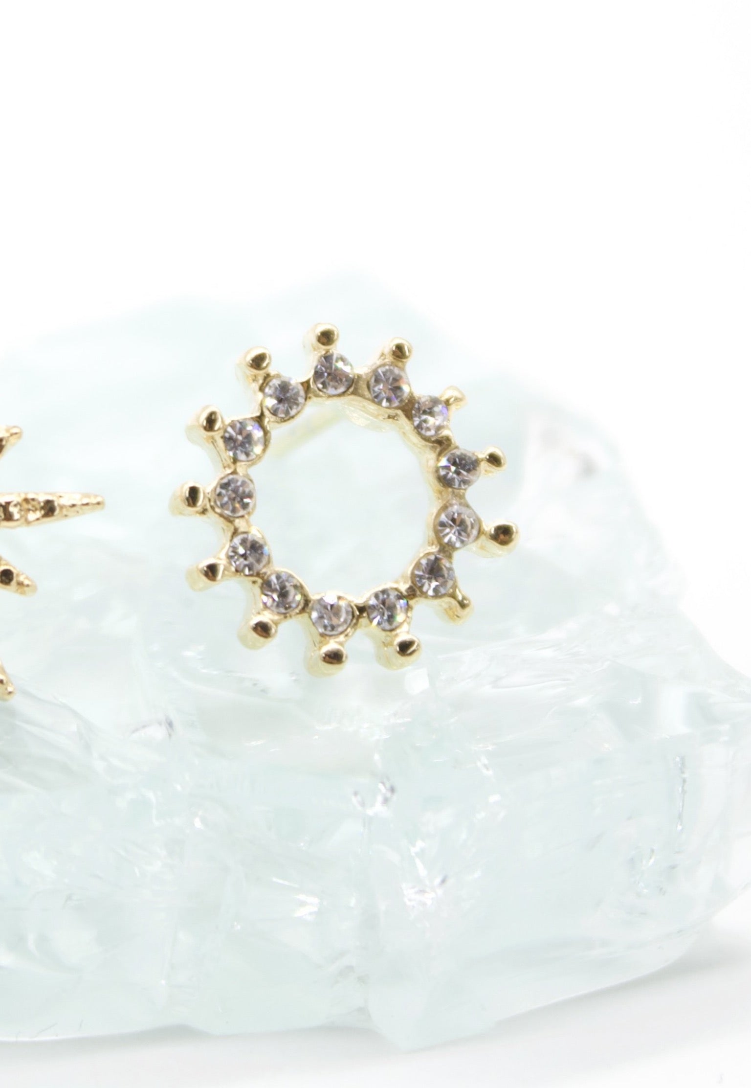 A gold sunburst stud earring adorned with sparkling cubic zirconia stones rests on a textured white surface.