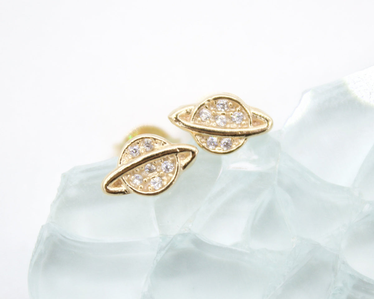 A pair of gold planet-shaped stud earrings featuring sparkling cubic zirconia details rests on a transparent crystal surface.