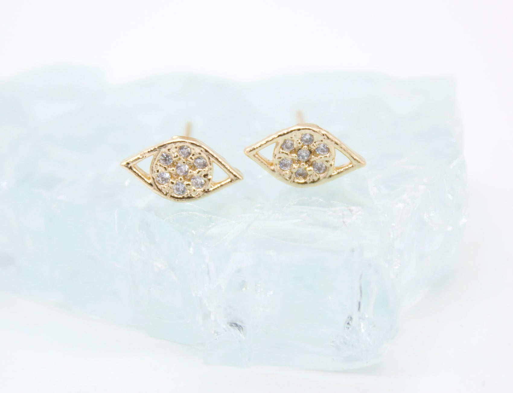 A pair of gold evil eye stud earrings featuring sparkling pave diamonds rests delicately on a piece of light blue sea glass.