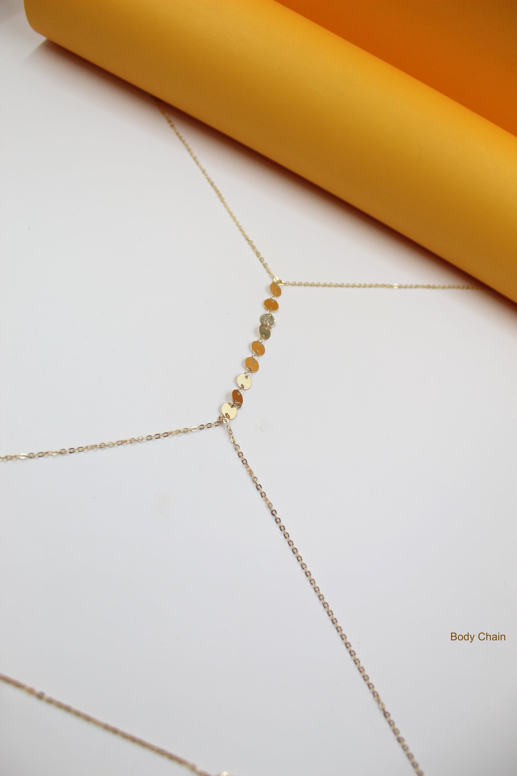 A delicate gold body chain featuring a vertical line of shimmering coin accents rests on a clean white surface against a bright yellow background.