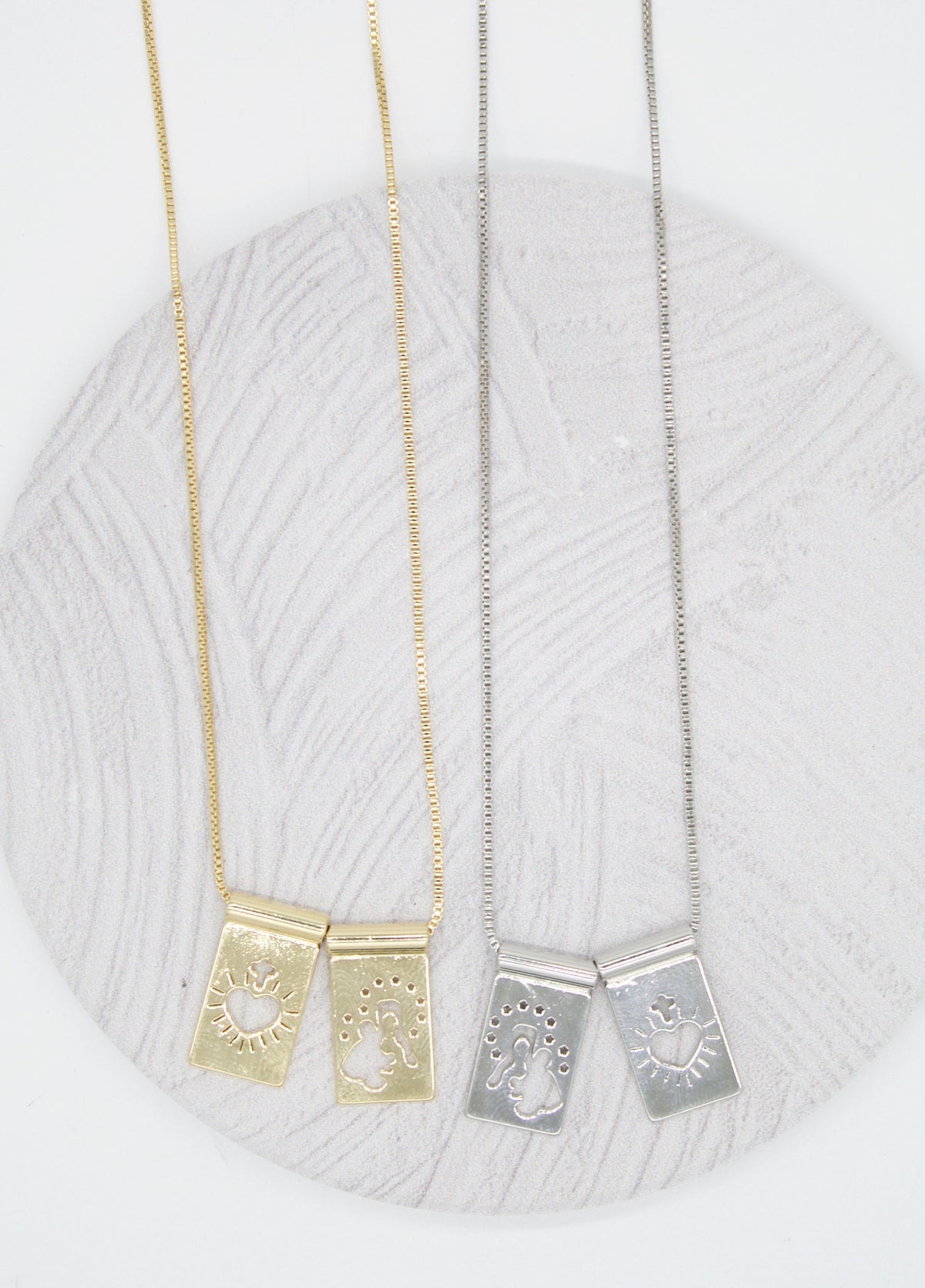 Two gold and two silver rectangular pendant necklaces with religious cut-out designs rest on a textured grey circular display.