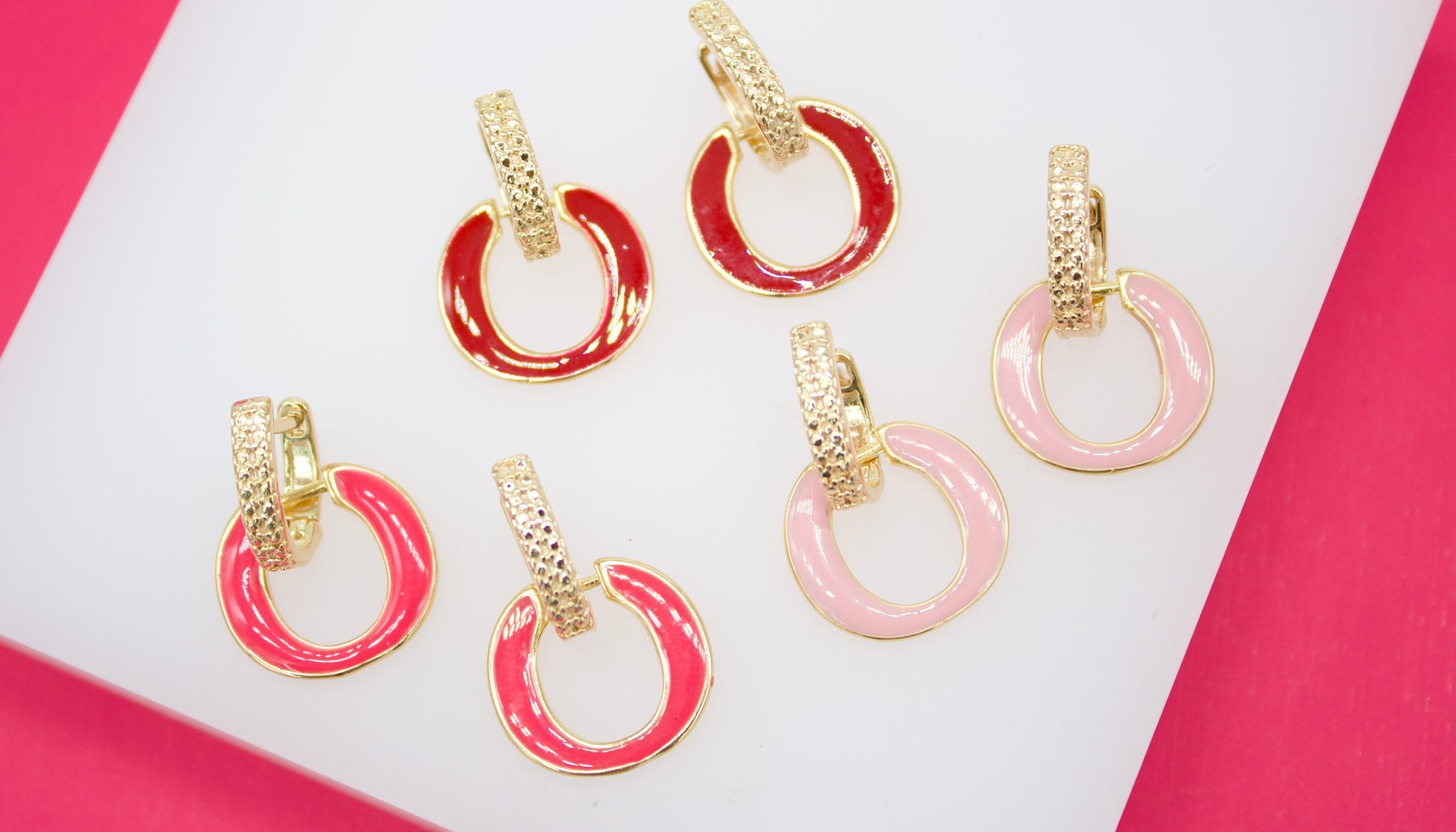 A collection of colorful enamel gold huggie hoop earrings displays red, magenta, and pink designs arranged on a white and bright pink display surface.