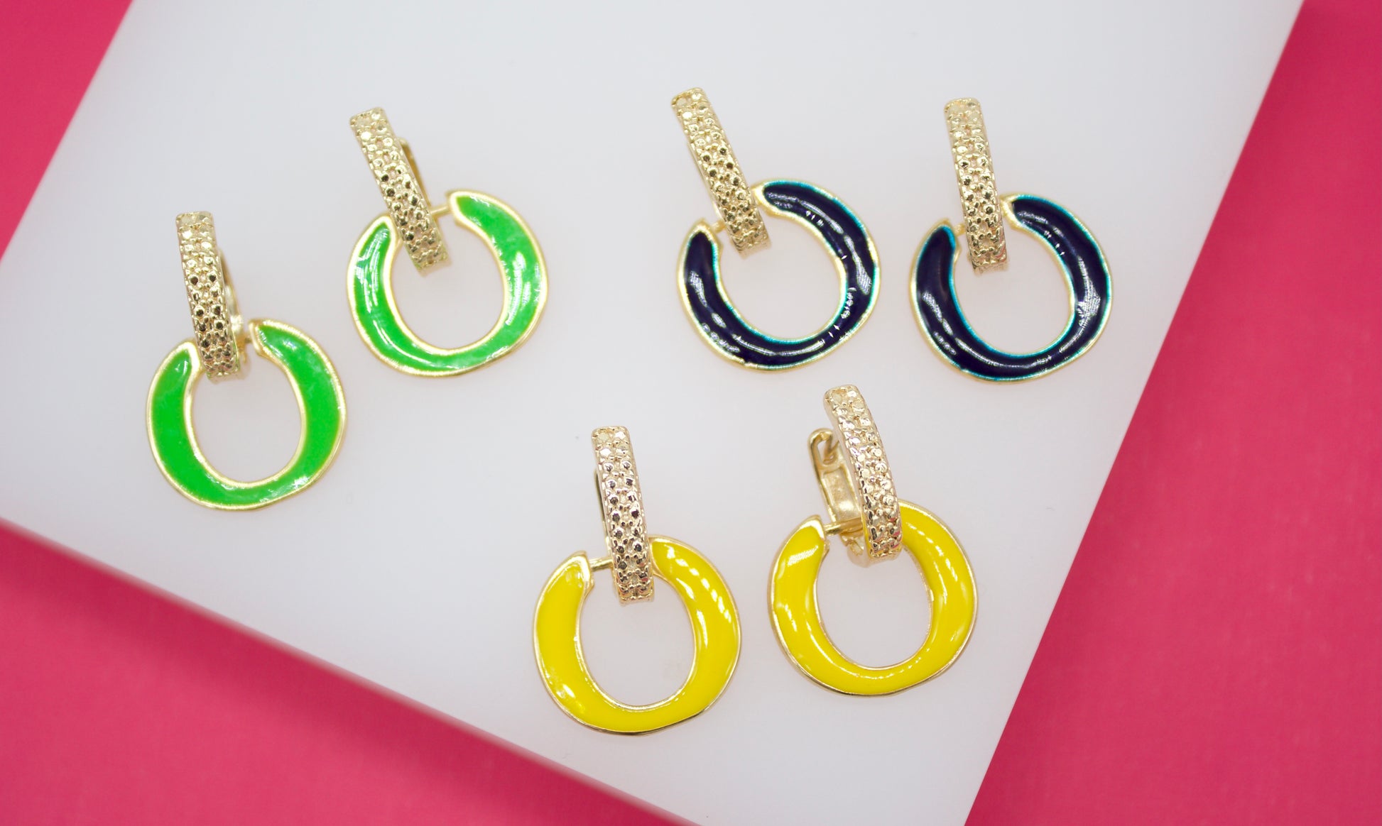 Three pairs of vibrant enameled hoop earrings in bright green, deep navy, and sunny yellow rest on a white display surface against a pink background.