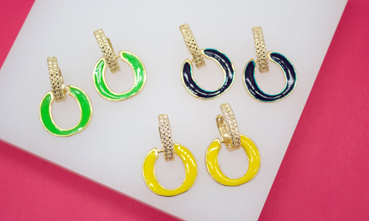 Three pairs of vibrant enameled hoop earrings in bright green, deep navy, and sunny yellow rest on a white display surface against a pink background.