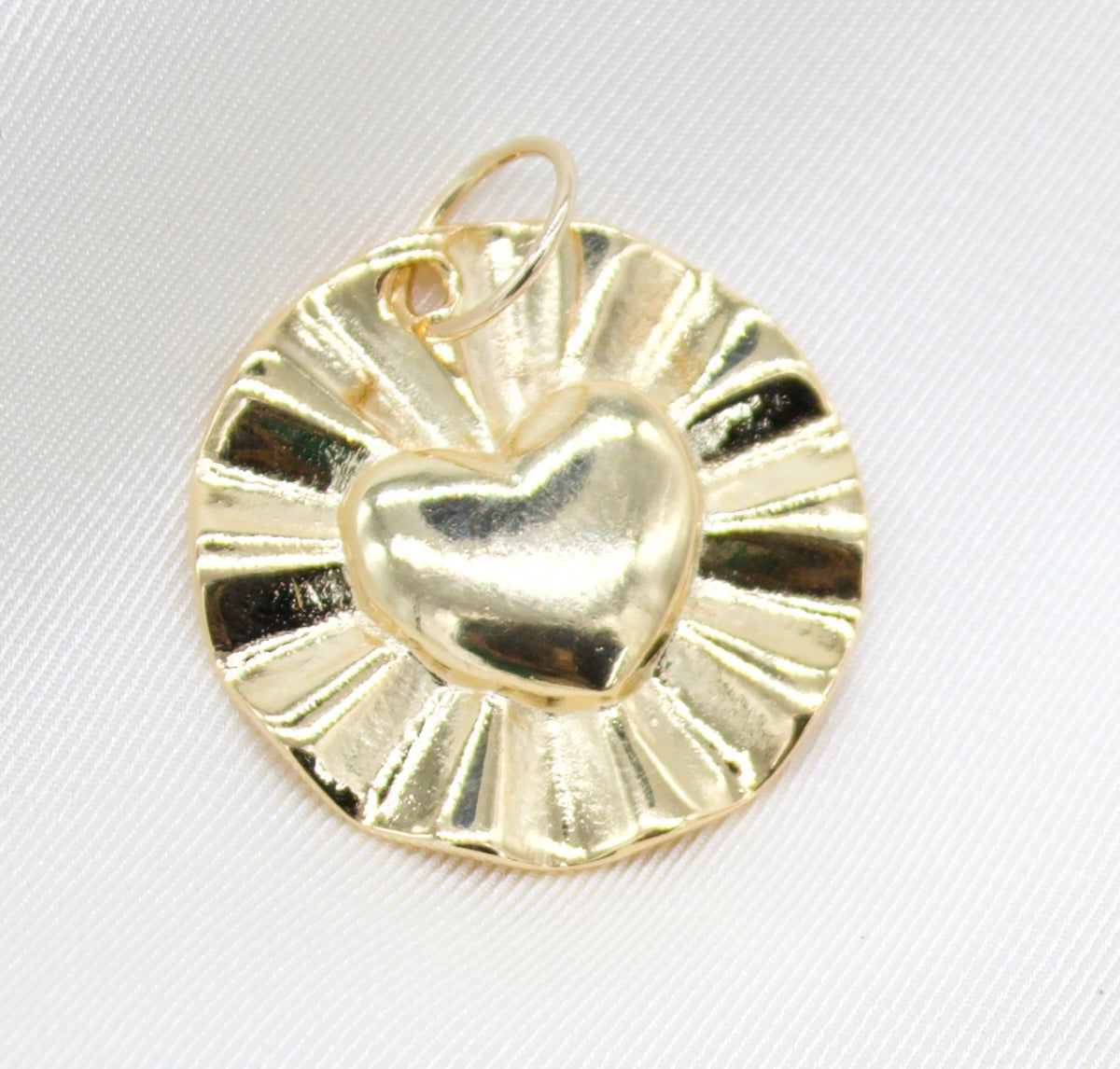 A shiny gold pendant features a raised puffy heart design centered on a round, sunburst-patterned disc resting against a white textured background.