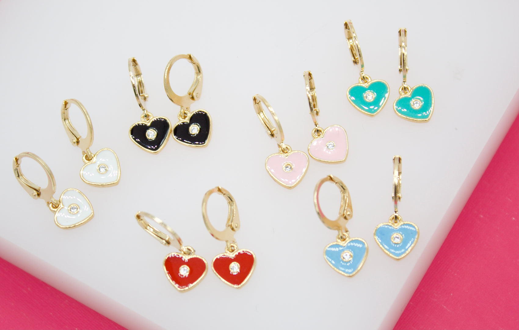Several pairs of colorful, heart-shaped huggie hoop earrings featuring center rhinestones are arranged on a white display surface.