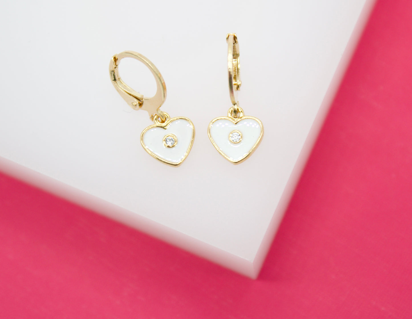 A pair of gold heart-shaped hoop earrings with white enamel and sparkling center stones rests on a white and pink display stand.