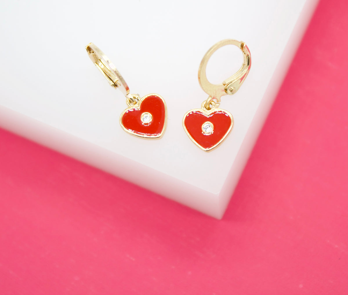 A pair of gold huggie hoop earrings featuring dangling red enamel heart charms with a small center crystal sits on a white and pink display.