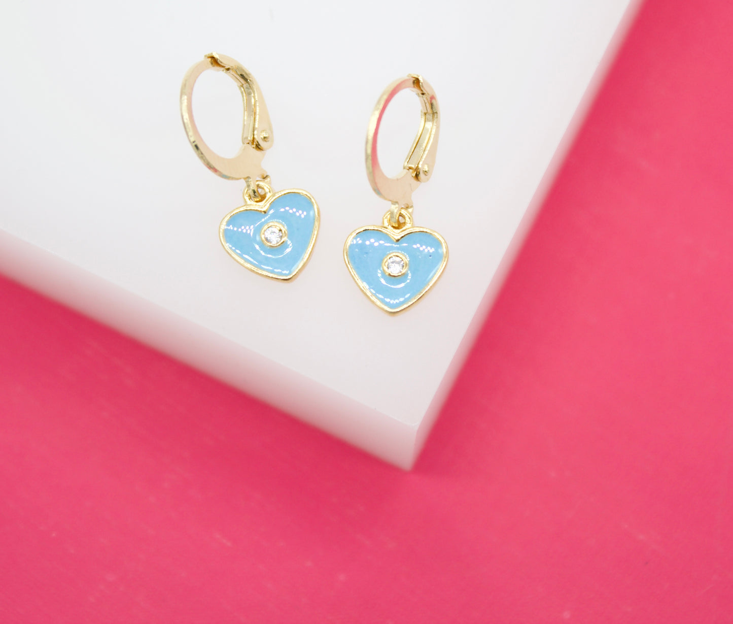 A pair of gold hoop earrings featuring light blue heart-shaped pendants with sparkling center stones rests on a white and pink display.