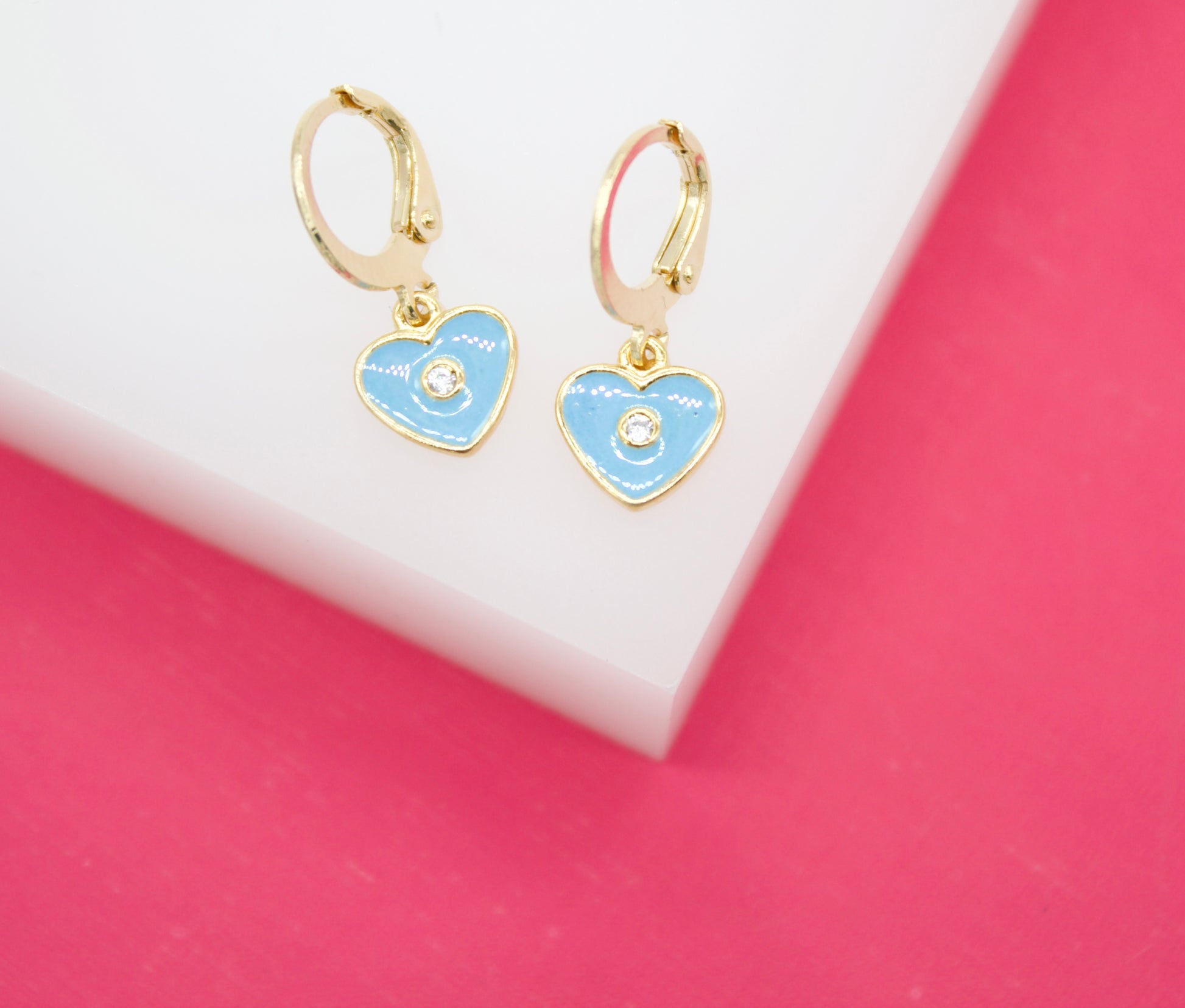 A pair of gold hoop earrings featuring light blue heart-shaped pendants with sparkling center stones rests on a white and pink display.