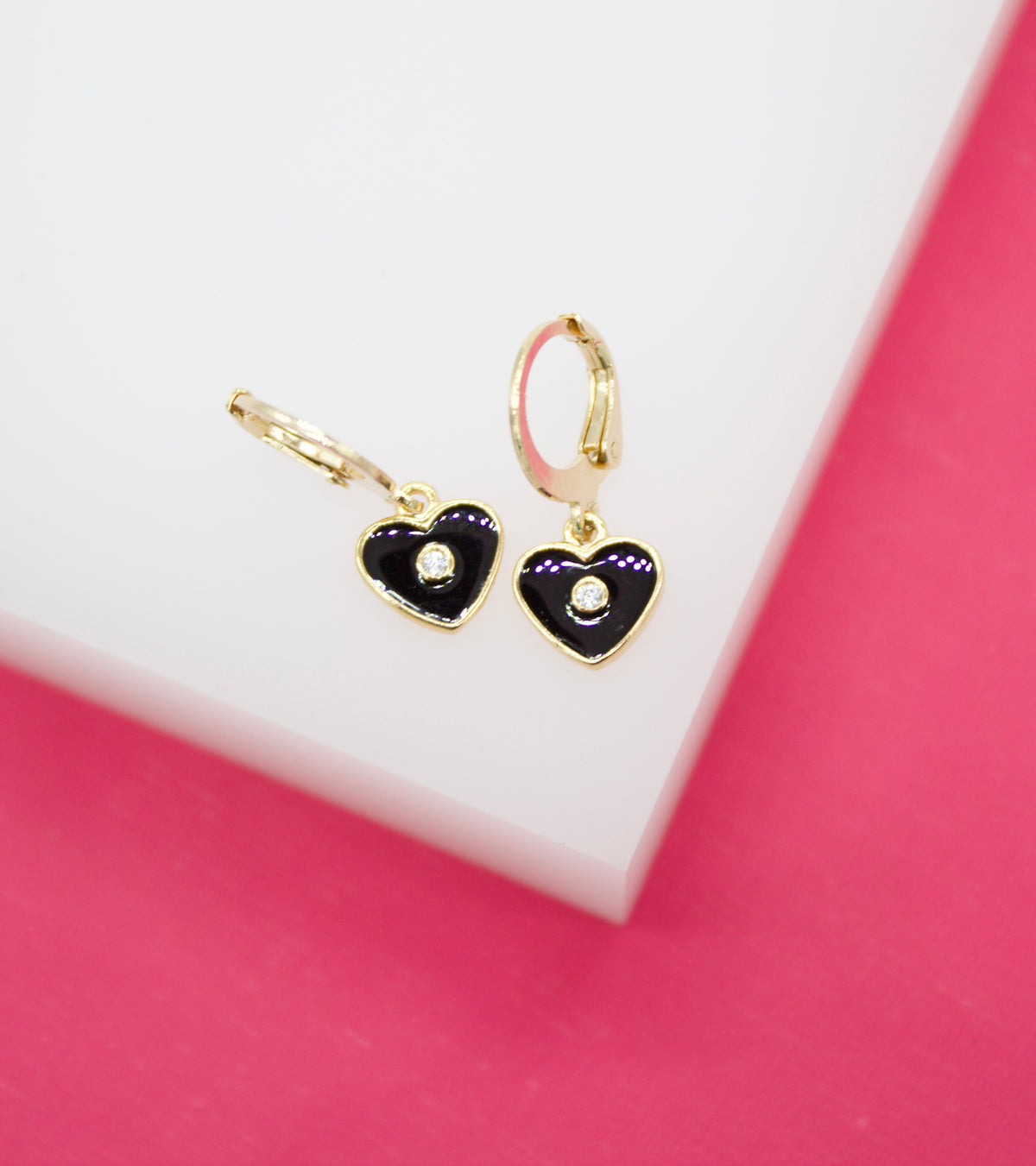 A pair of gold huggie hoop earrings featuring black enamel heart charms with center crystals rests on a white and pink display surface.