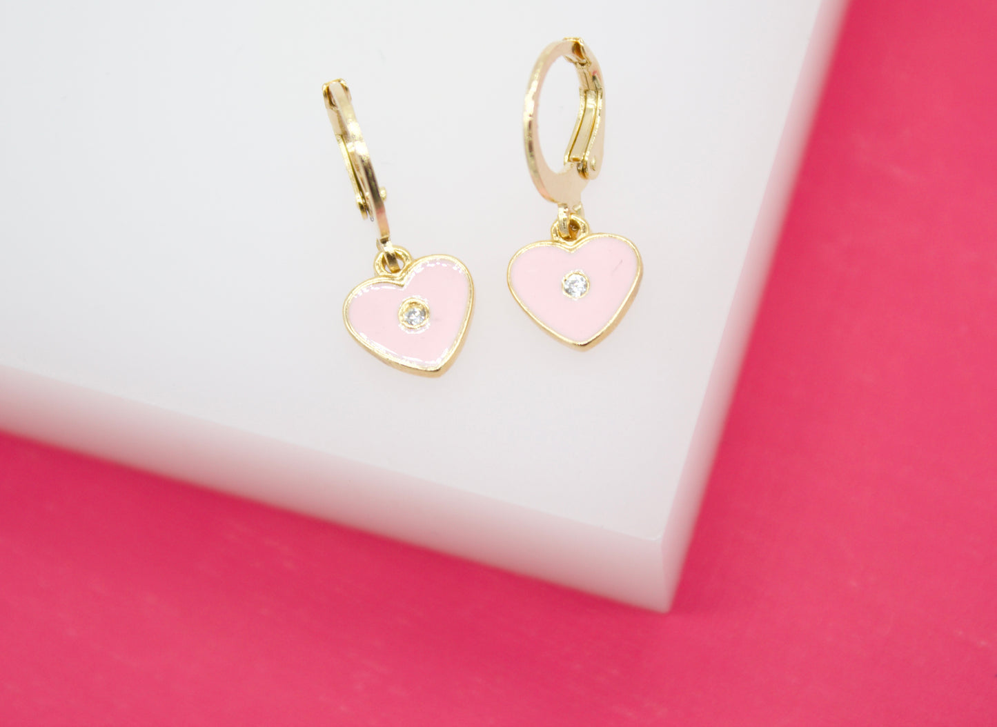 Two gold hoop earrings featuring pink enamel heart charms with center crystals rest on a white display block against a vibrant pink background.