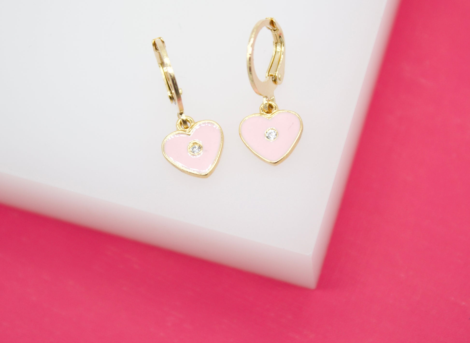 Two gold hoop earrings featuring pink enamel heart charms with center crystals rest on a white display block against a vibrant pink background.