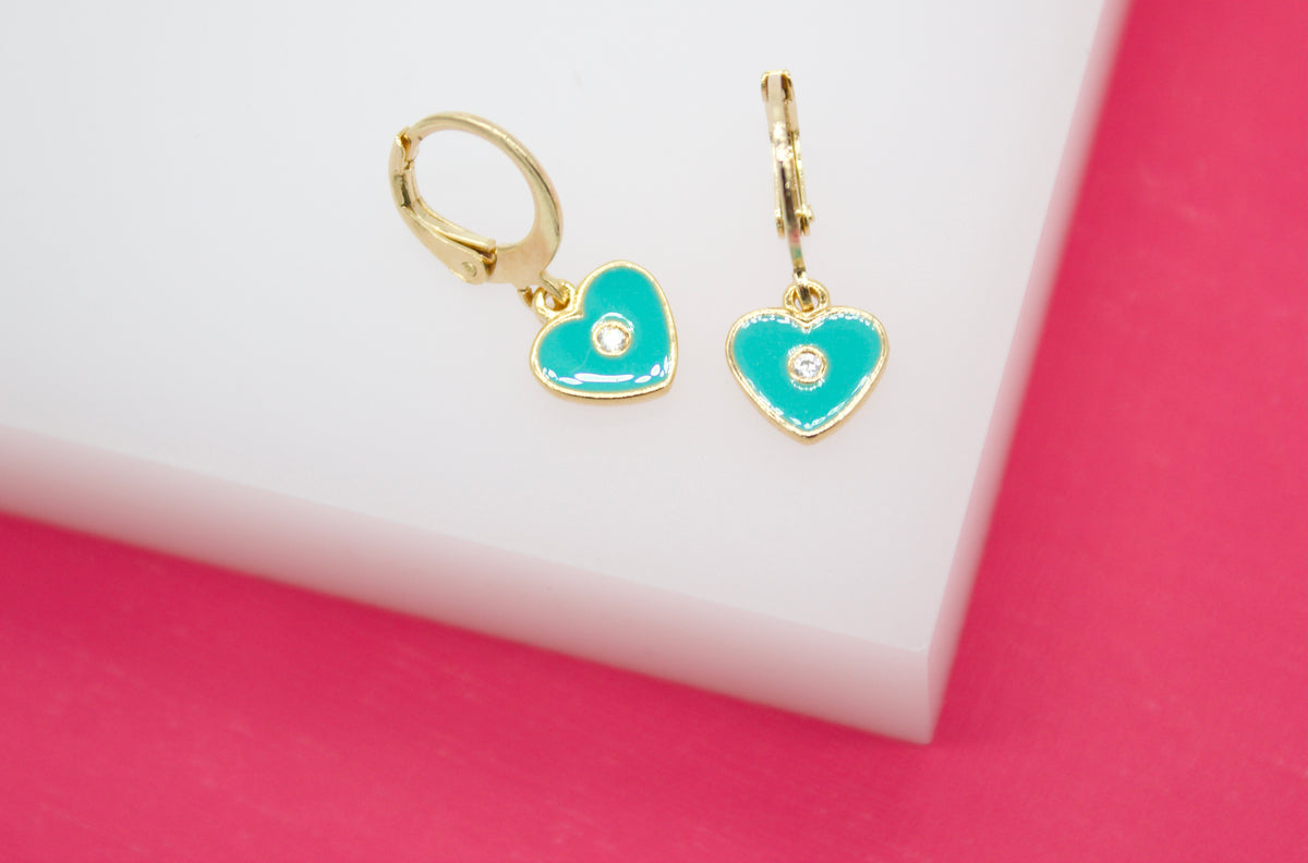 A pair of gold huggie hoop earrings featuring enamel turquoise heart charms with central diamond accents rests on a white display block against a vibrant pink background.