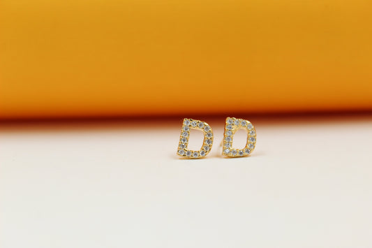 A pair of gold initial D stud earrings sparkles against a light background with an orange backdrop.