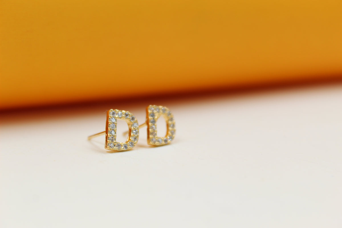 A pair of gold initial D stud earrings encrusted with small sparkling diamonds rest on a light surface against an orange backdrop.