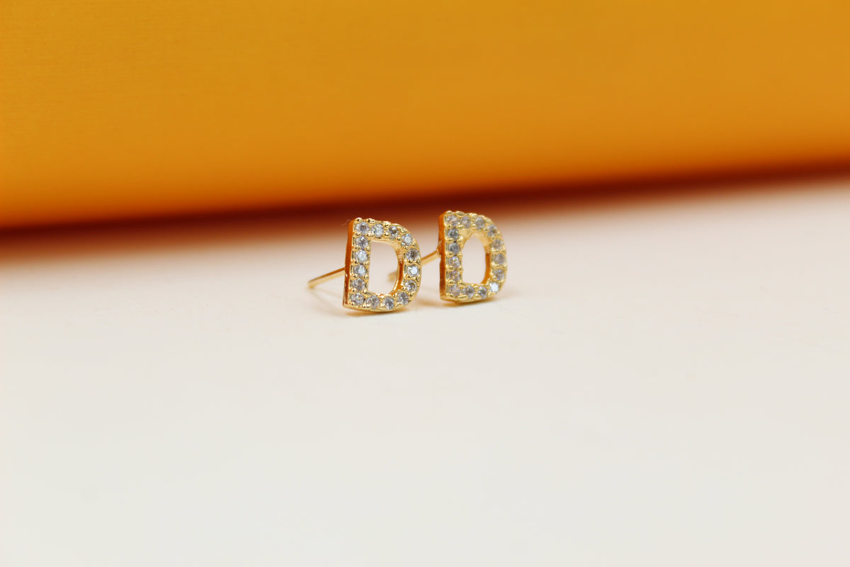 A pair of gold initial D stud earrings sparkles against a light-colored background.