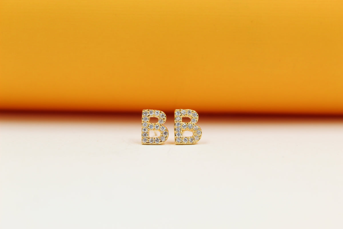 A pair of gold initial B earrings encrusted with sparkling cubic zirconia stones rests against a vibrant orange and white background.