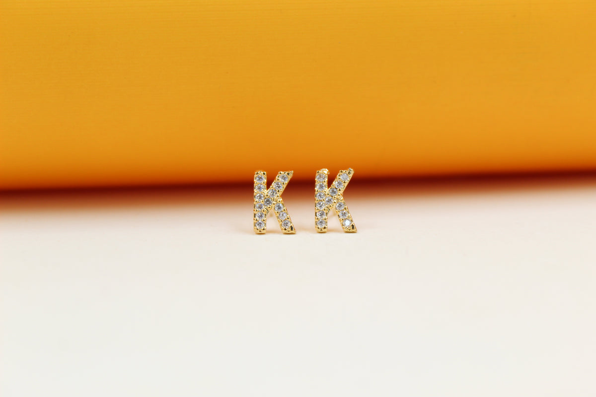 A pair of gold initial K earrings encrusted with sparkling cubic zirconia crystals rests on a light surface against an orange backdrop.