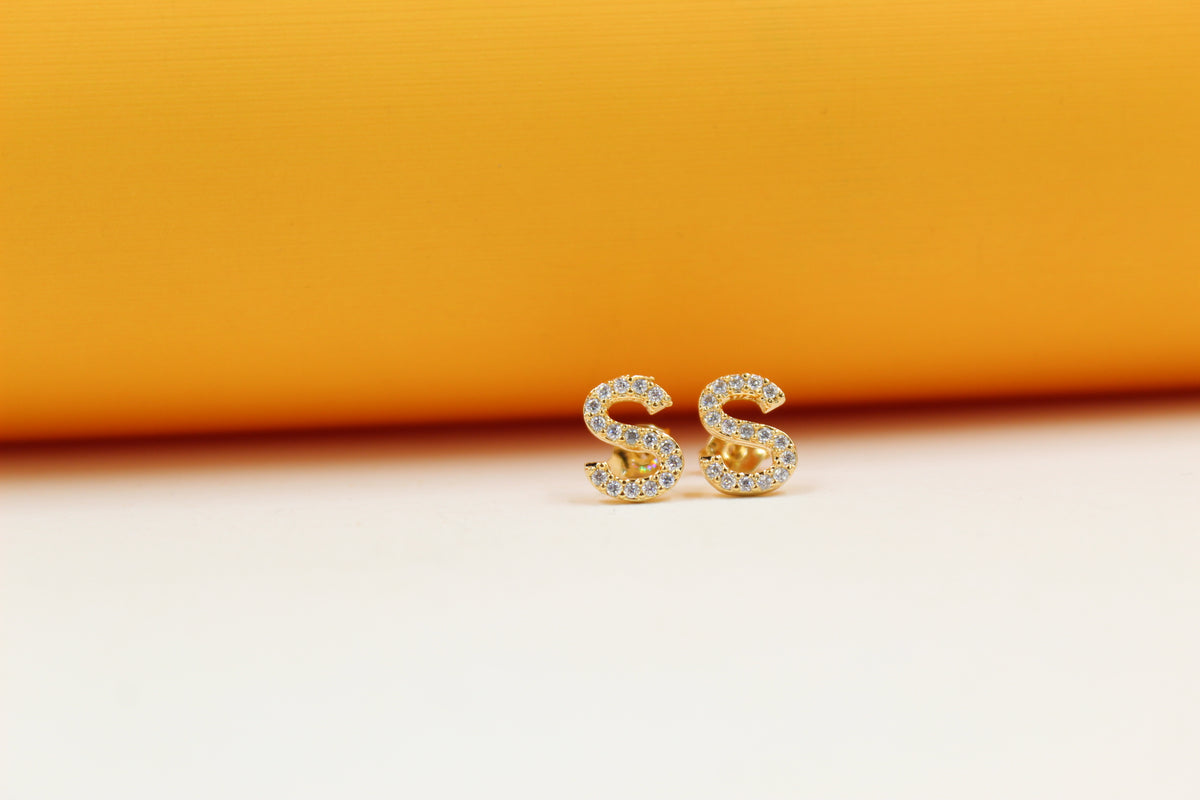 A pair of gold letter S stud earrings embellished with sparkling cubic zirconia stones rests against a warm yellow and white background.