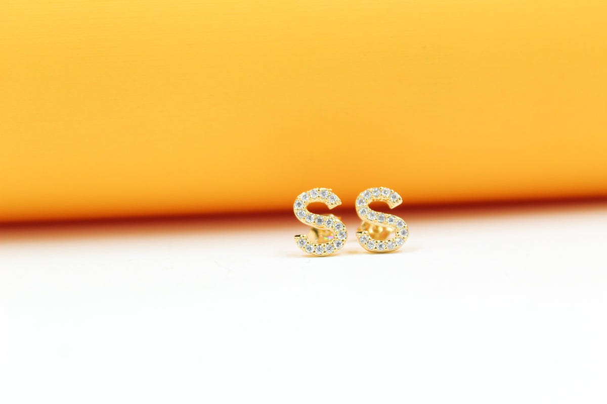 A pair of gold initial S earrings encrusted with sparkling cubic zirconia rest against a bright yellow background.
