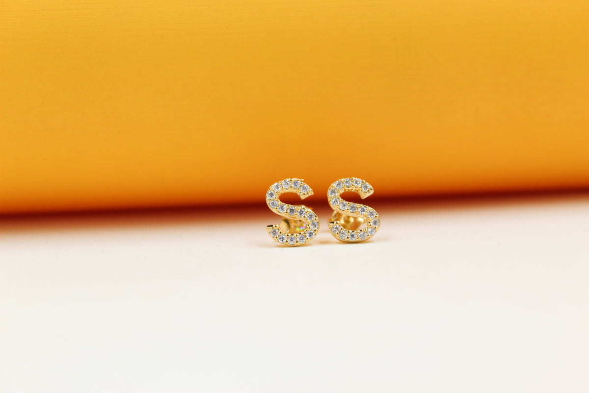 A pair of gold letter S stud earrings encrusted with sparkling cubic zirconia stones rests against a vibrant orange and white background.