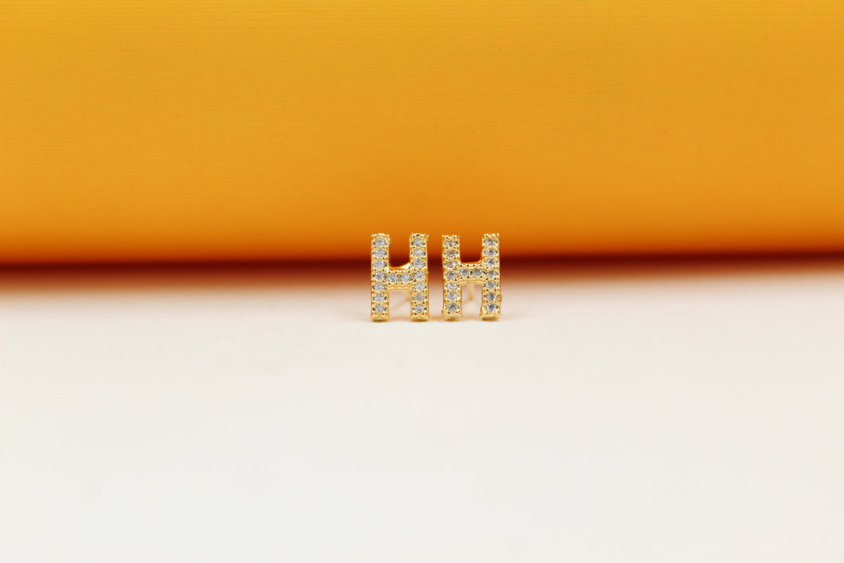 A pair of gold initial H earrings embedded with small cubic zirconia stones rests against a vibrant orange and white background.