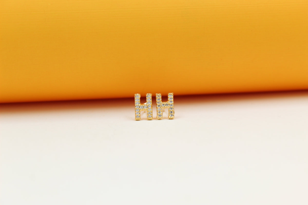 A pair of gold initial H earrings embedded with tiny diamonds sits on a white surface against a yellow background.