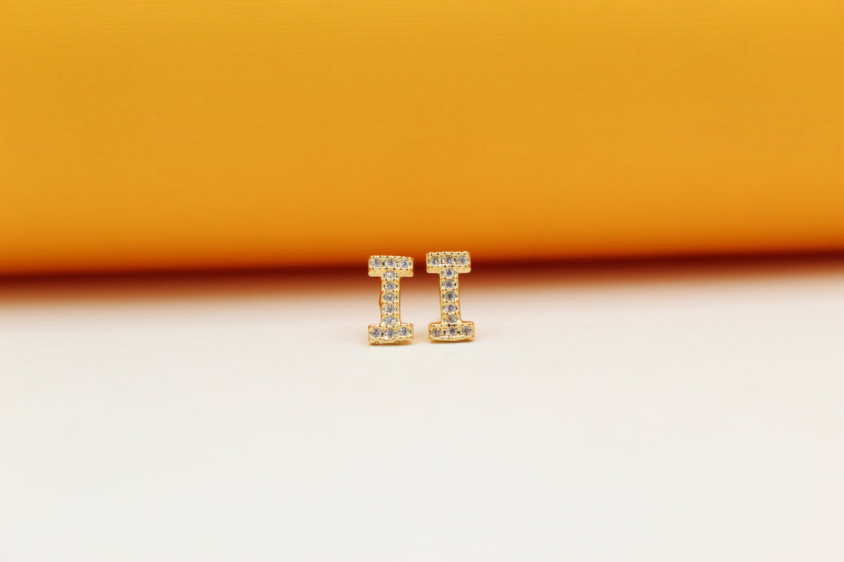 A pair of gold initial I stud earrings encrusted with sparkling cubic zirconia stones rests upright on a neutral surface against a vibrant orange and white background.