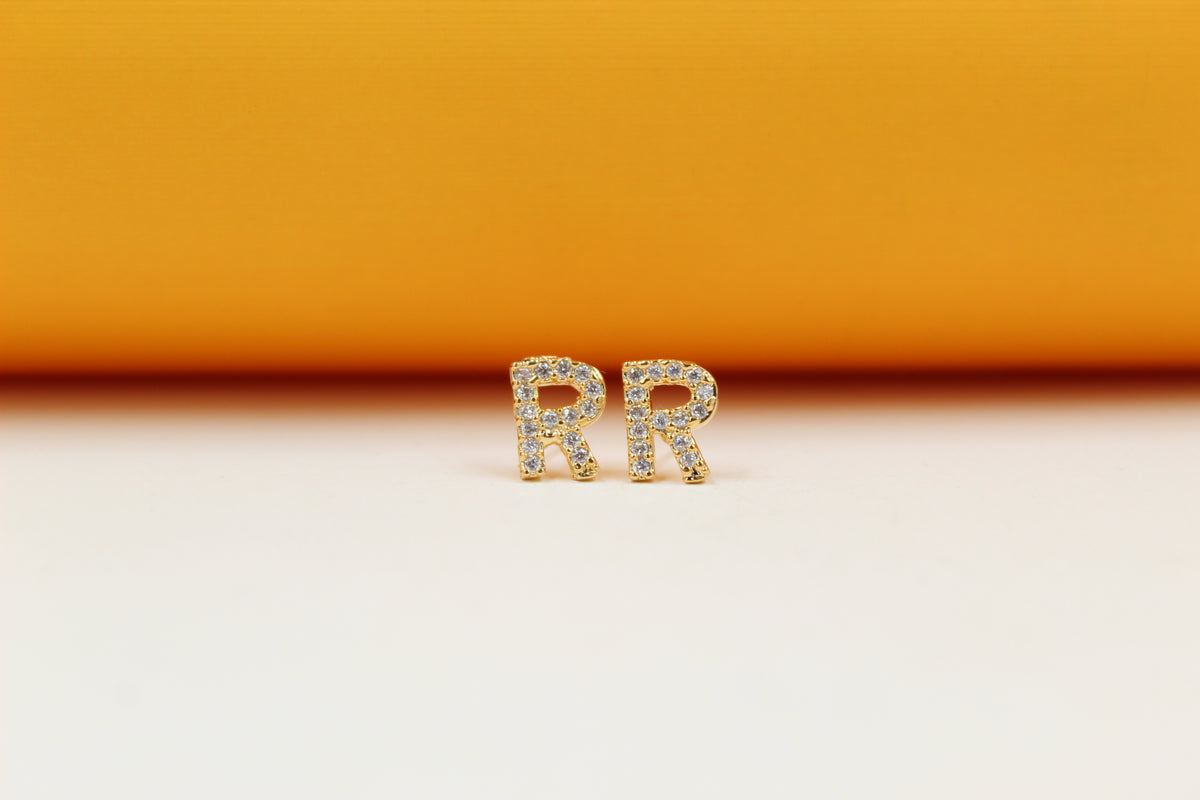 A pair of gold initial R earrings studded with small sparkling diamonds sit on a neutral surface against an orange background.