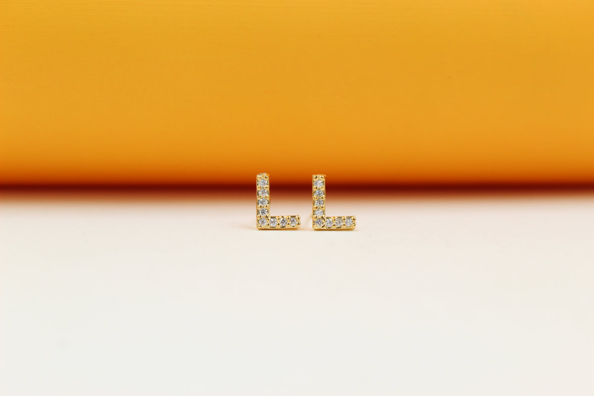 Two gold letter L initial stud earrings embedded with sparkling cubic zirconia stones rest side by side against a warm gradient background.