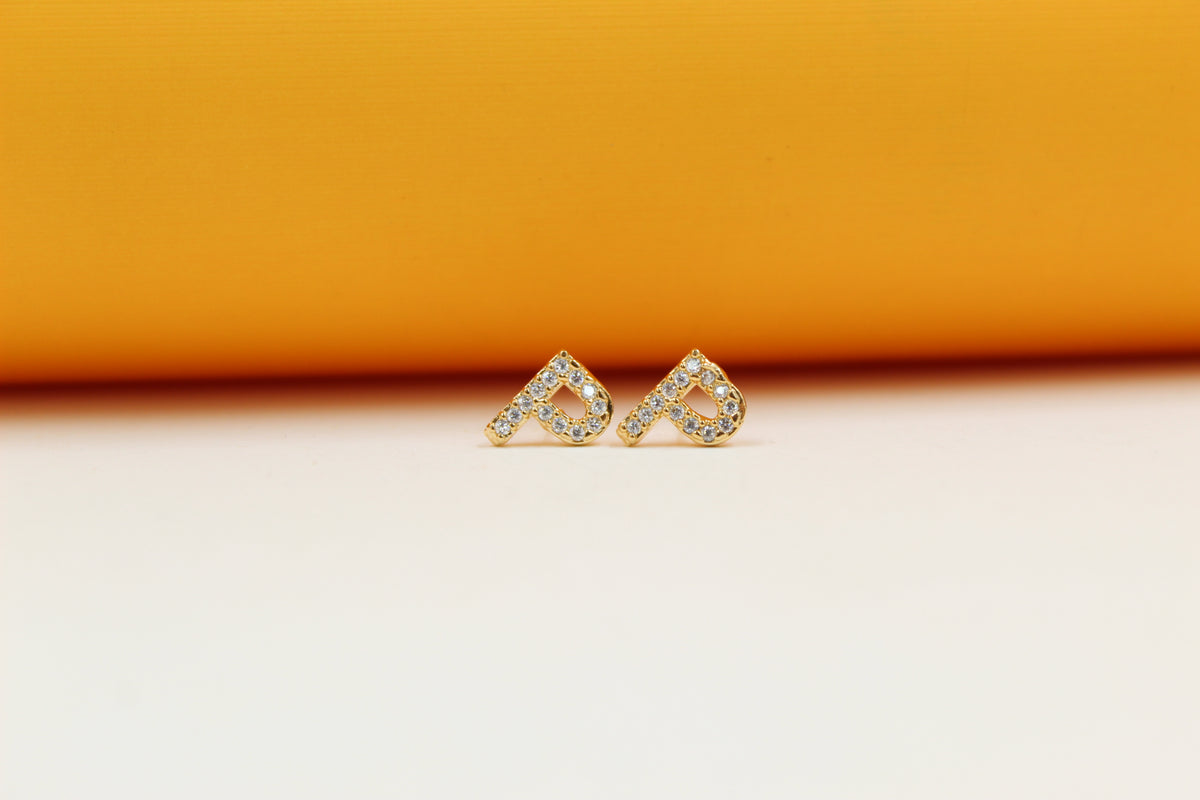 A pair of gold initial P stud earrings encrusted with sparkling cubic zirconia stones rests against a vibrant orange and cream background.