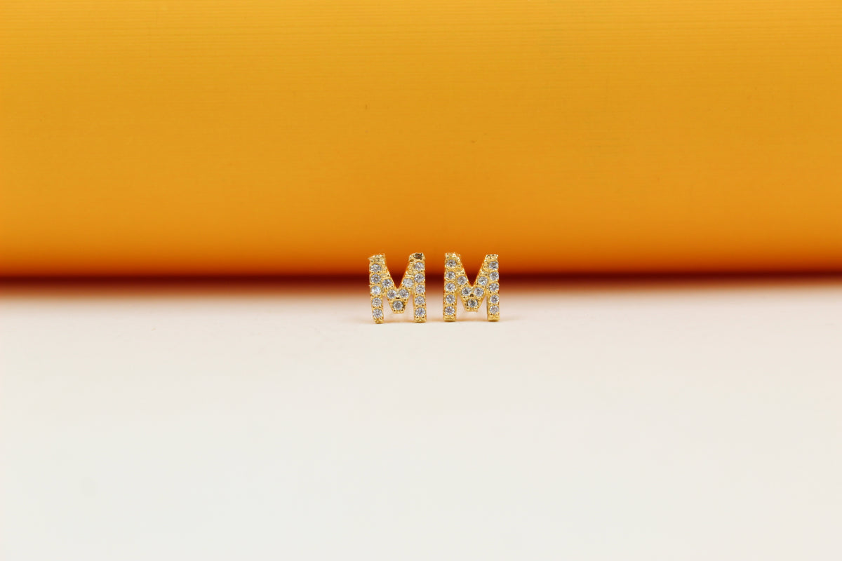 A pair of gold initial M stud earrings sparkle against a vibrant orange and white background.
