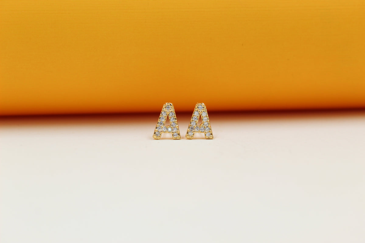 A pair of gold initial letter A stud earrings encrusted with small clear cubic zirconia stones rests against a cream and orange background.