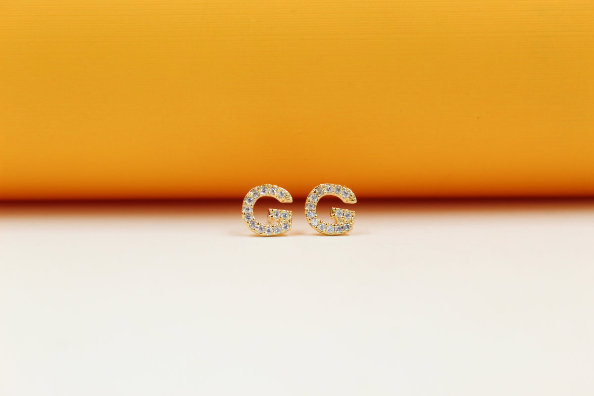 A pair of gold initial G earrings embellished with shimmering white crystals sits on a light surface against a bright orange background.