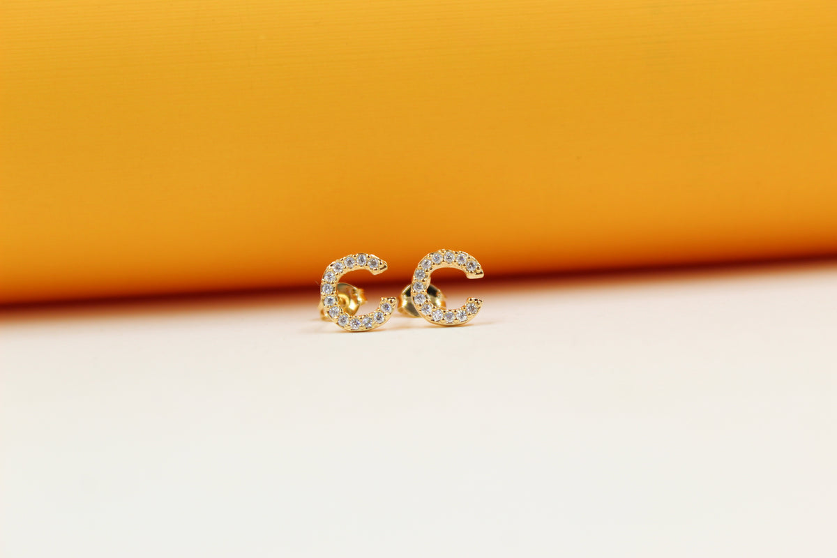 Two gold initial C earrings studded with small white crystals rest on a white surface against an orange backdrop.