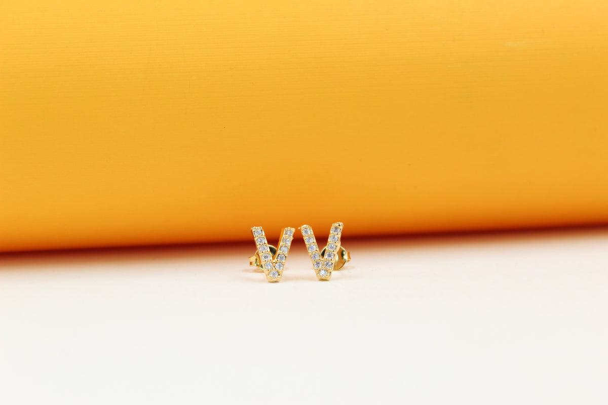 A pair of gold letter V earrings encrusted with sparkling cubic zirconia stones rests against a vibrant yellow and white background.