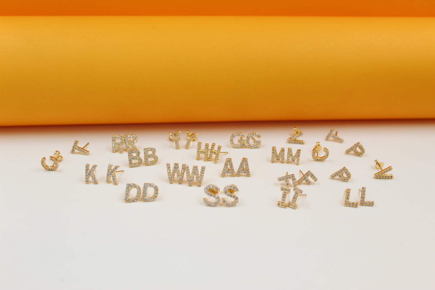 A collection of glittering pavé crystal gold letter initial earrings rests on a smooth surface against a bright orange and white backdrop.
