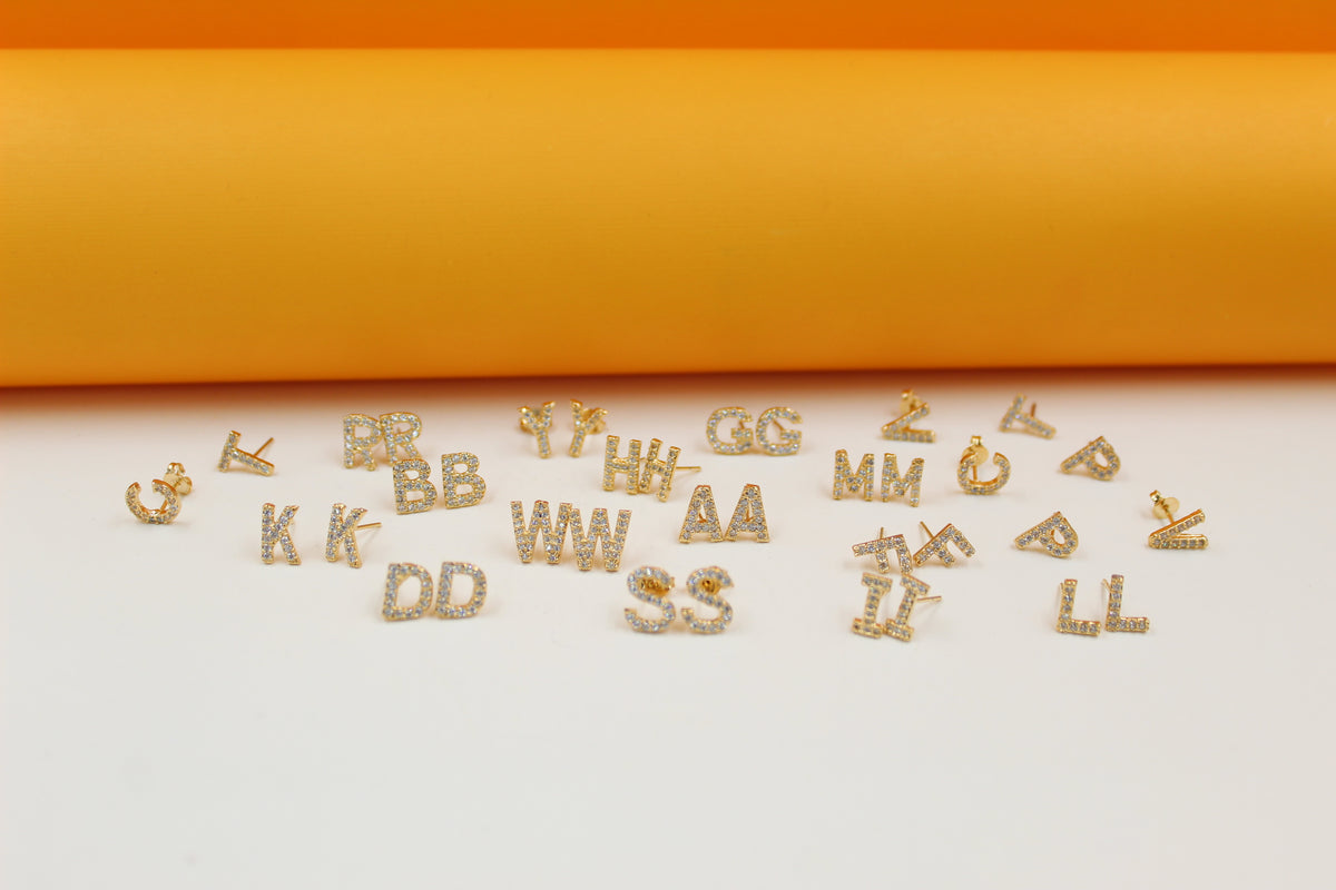 A collection of glittering pavé crystal gold letter initial earrings rests on a smooth surface against a bright orange and white backdrop.