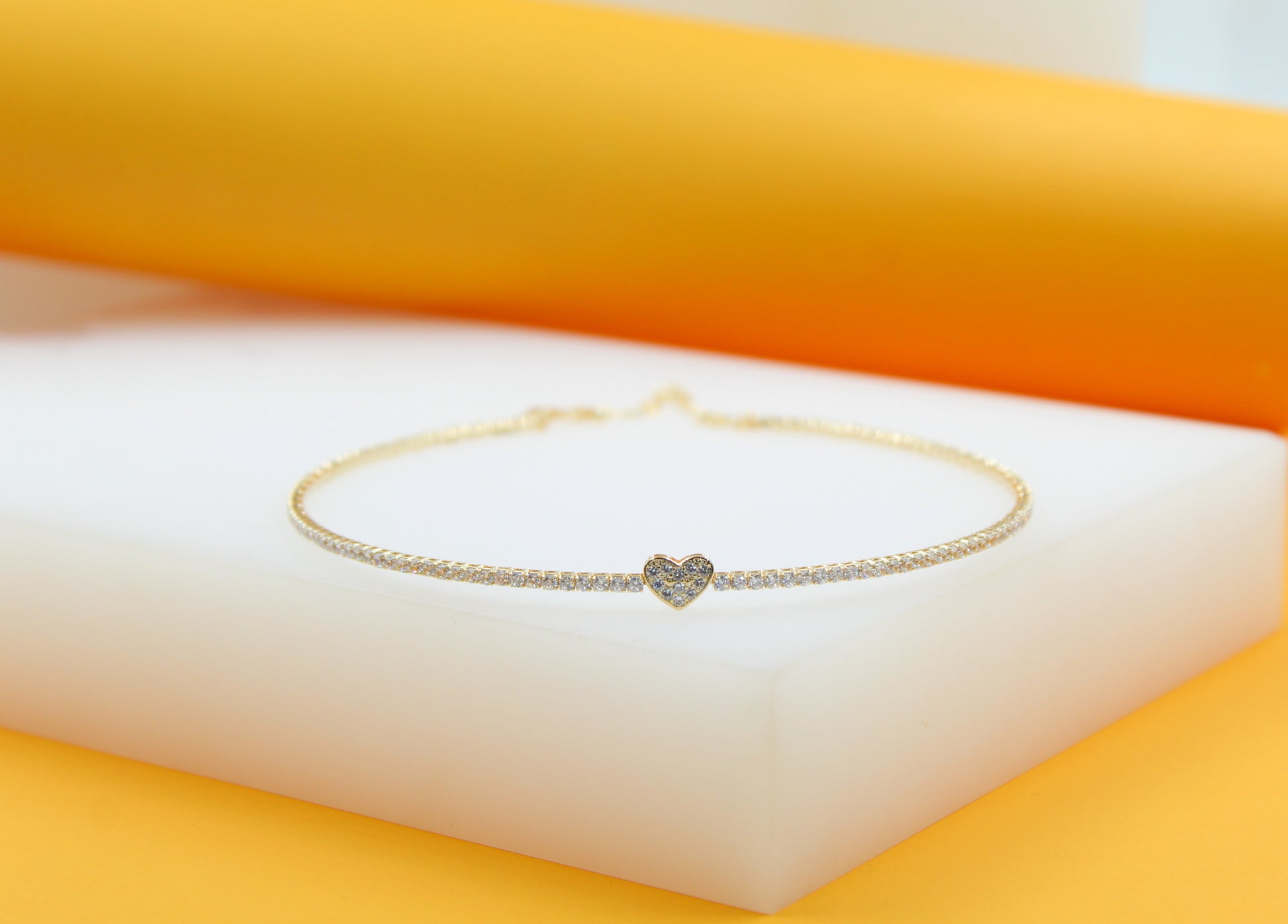 A delicate gold tennis necklace featuring a sparkling diamond heart pendant rests on a white display block against an orange backdrop.