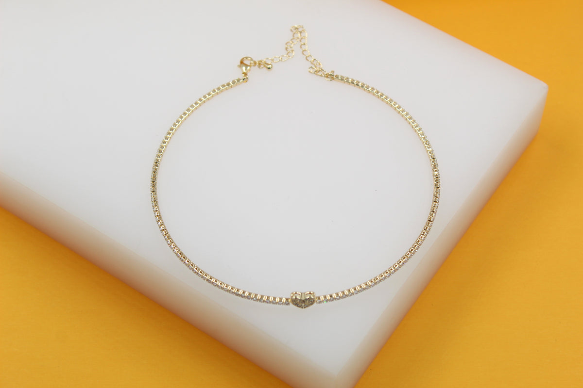 A gold tennis necklace featuring a delicate crystal-embellished heart pendant rests on a white display surface against a vibrant yellow background.